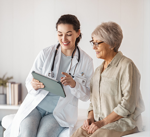 Doctor smiling and showing information on a tablet to a senior woman in a clinic setting.